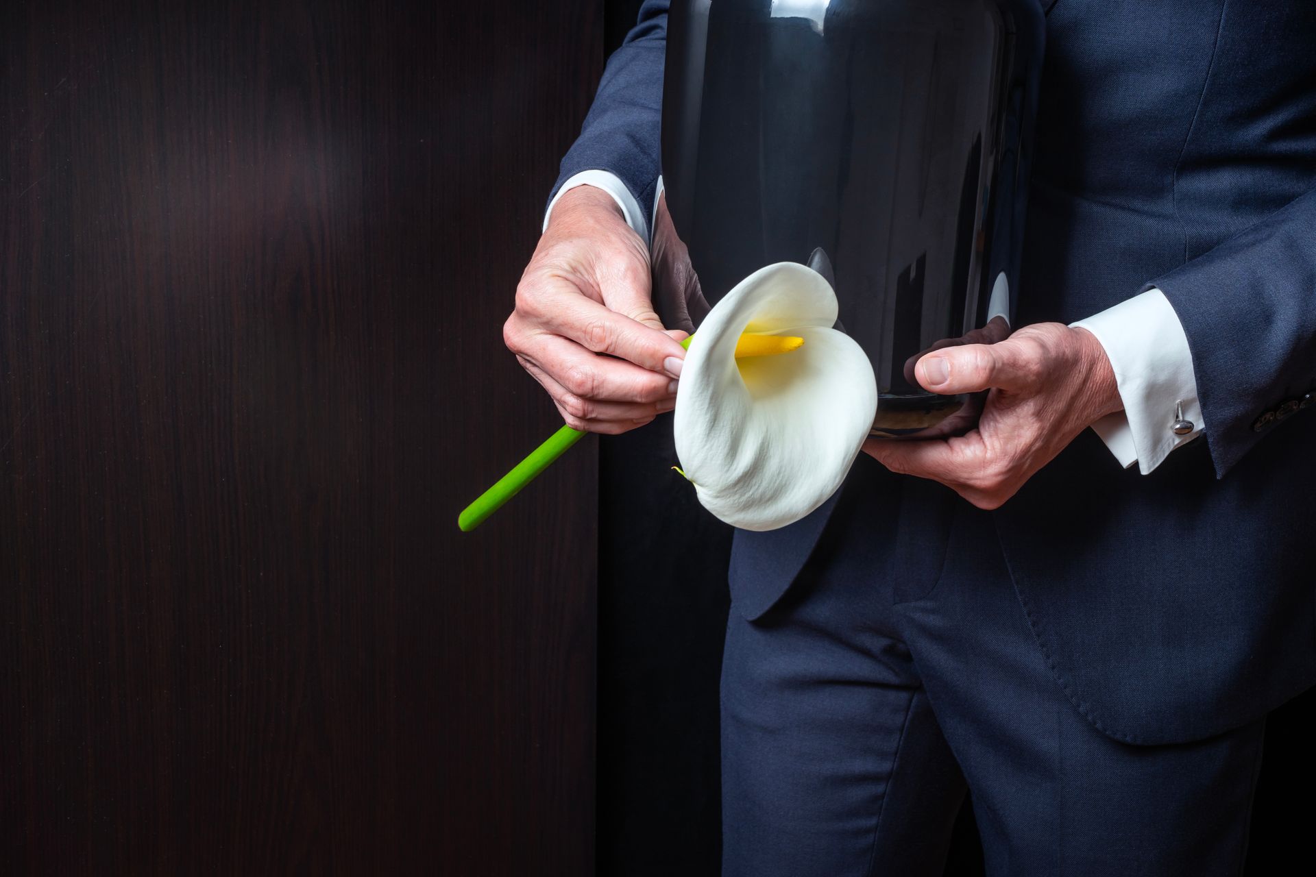 A person in a dark suit holds a smooth, black cremation urn adorned with a white calla lily against a dark background.