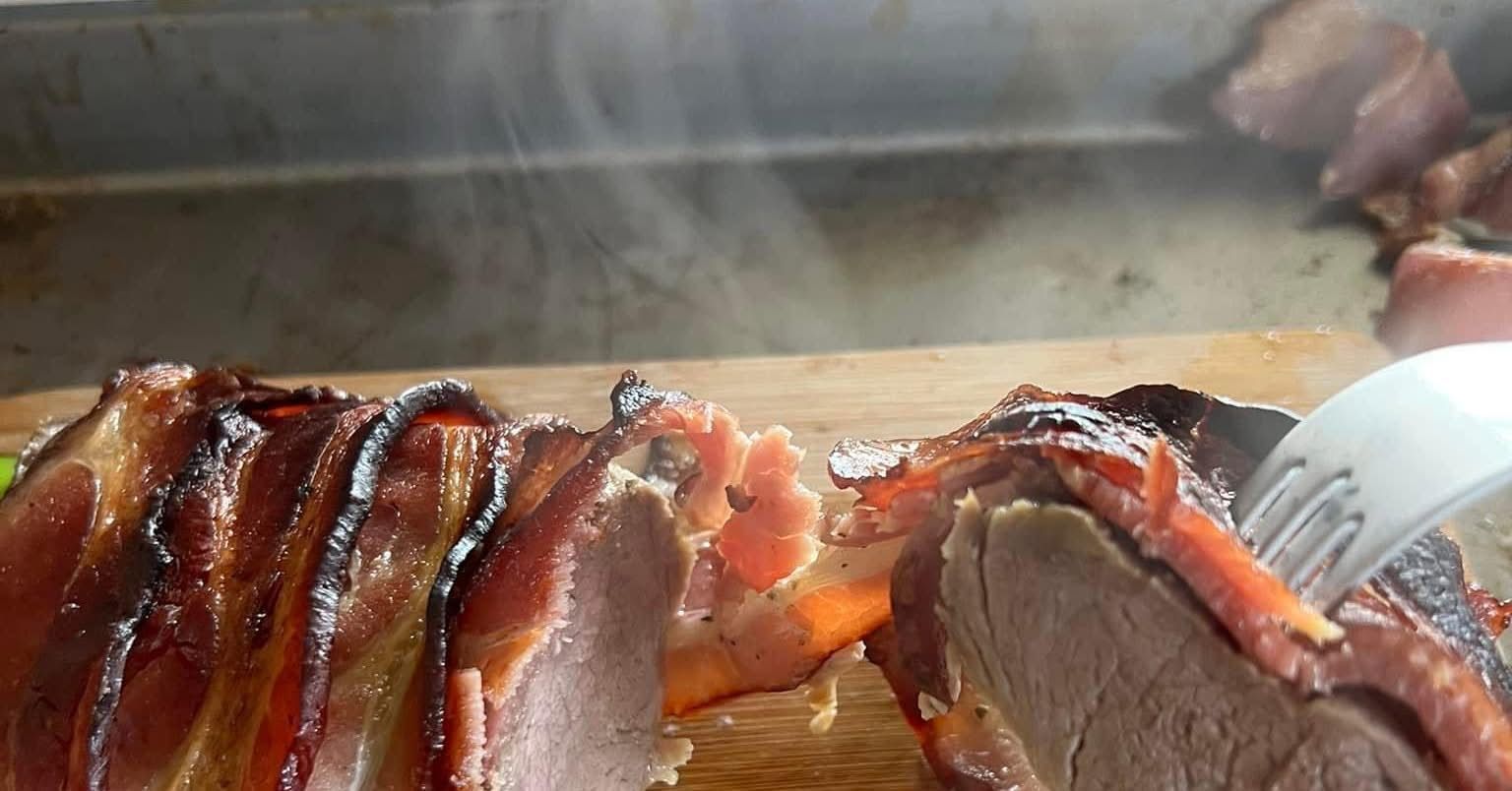 Slices of glazed smoked meat on a grill, with a smoky cooking surface and serving tongs nearby