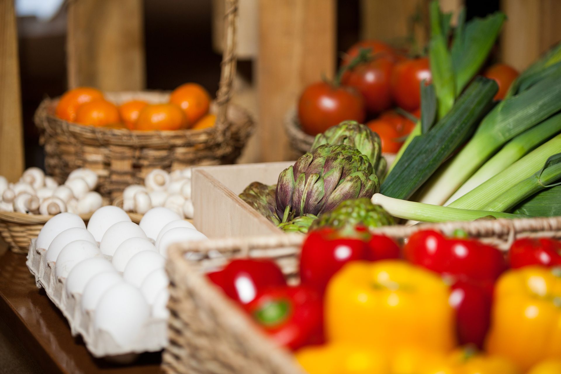 Baskets of fresh eggs, tomatoes, peppers, leeks, artichokes, and oranges at a market stall