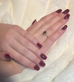Hands with burgundy-colored nail polish, one hand resting on the other, wearing a gold ring with a red gemstone.