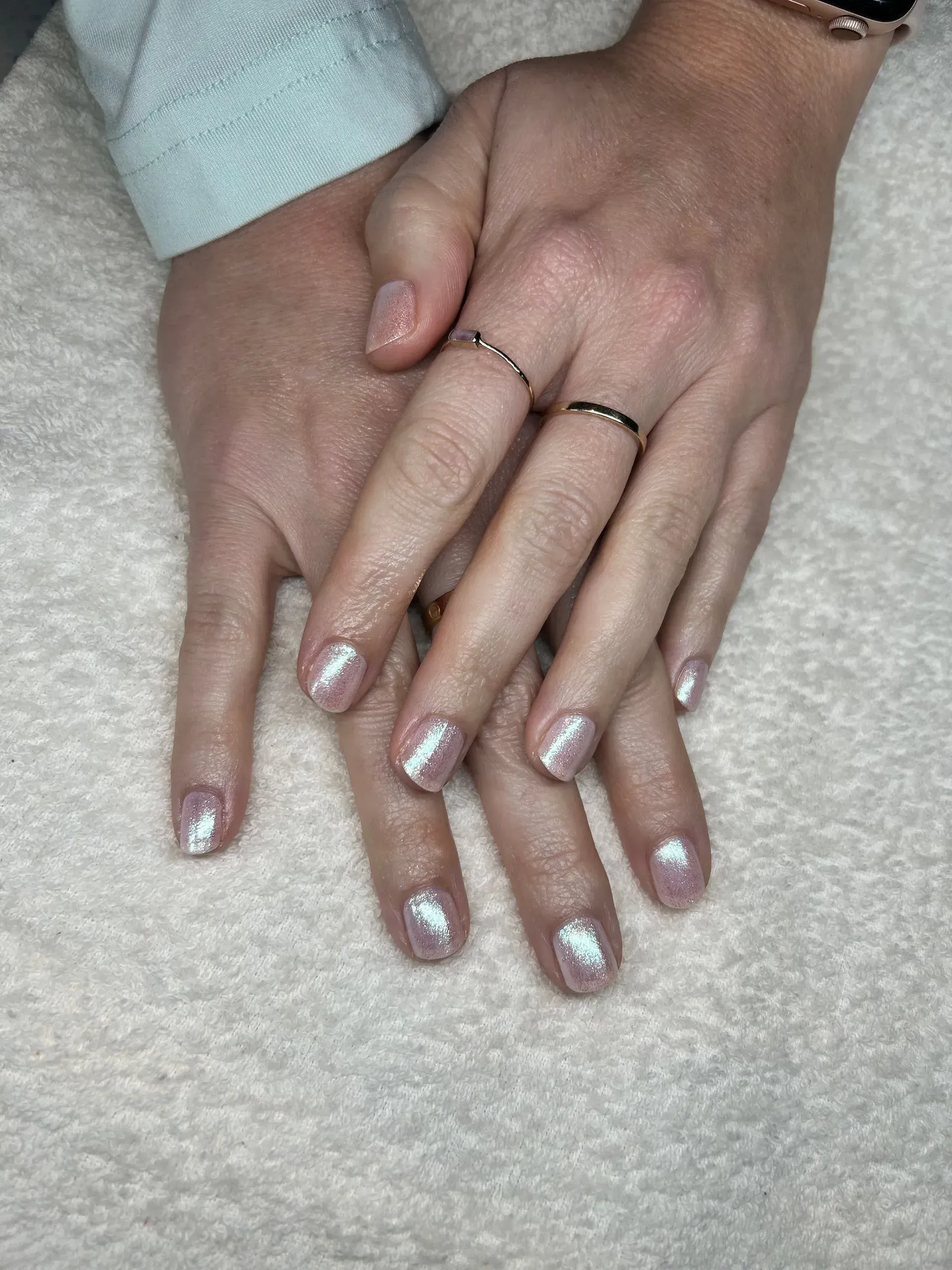 Hands with shimmering light pink nail polish, wearing rings, resting on a white surface. Hands with shimmering light pink nail polish, wearing rings, resting on a white surface.