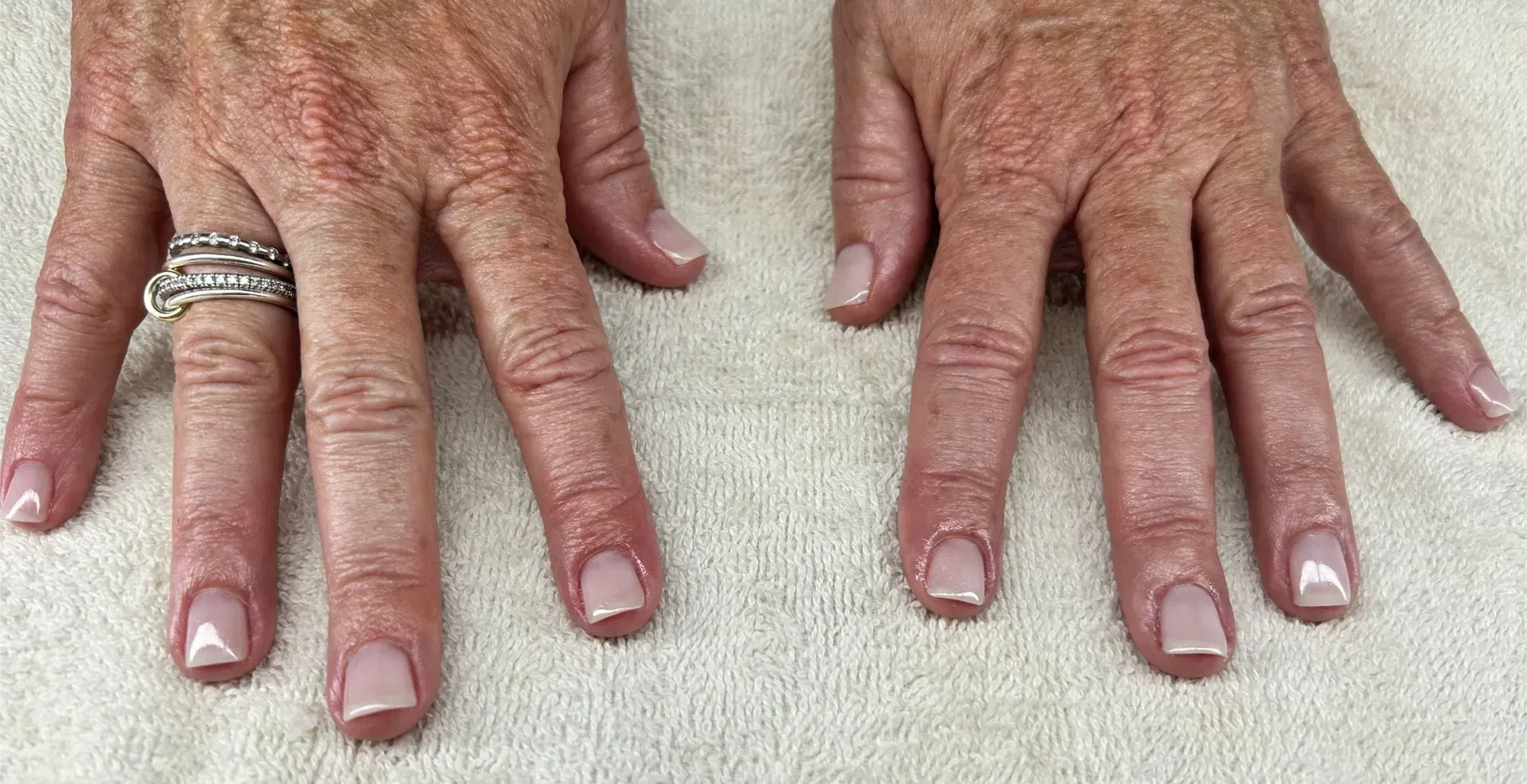Two hands with light pink nail polish, a ring on the left ring finger, resting on a white surface.