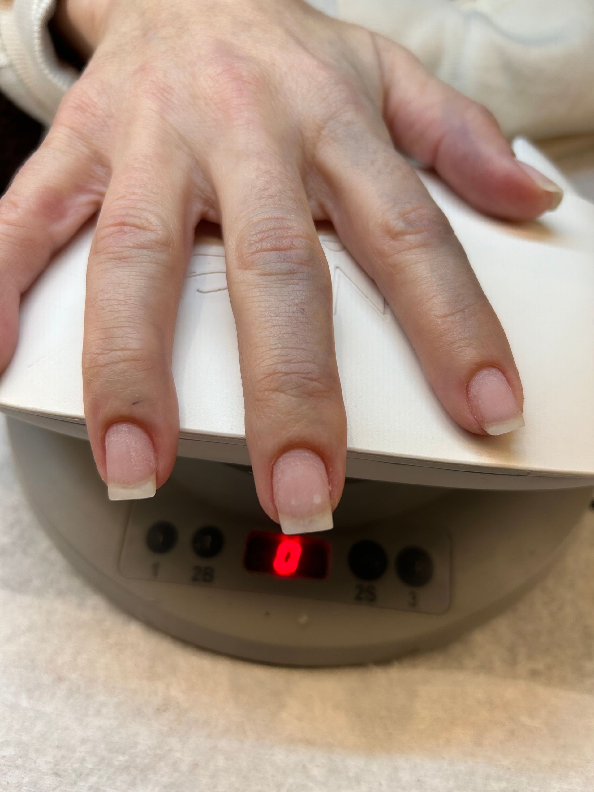 Hand with French-tipped nails on a manicure machine; digital display shows a red light.