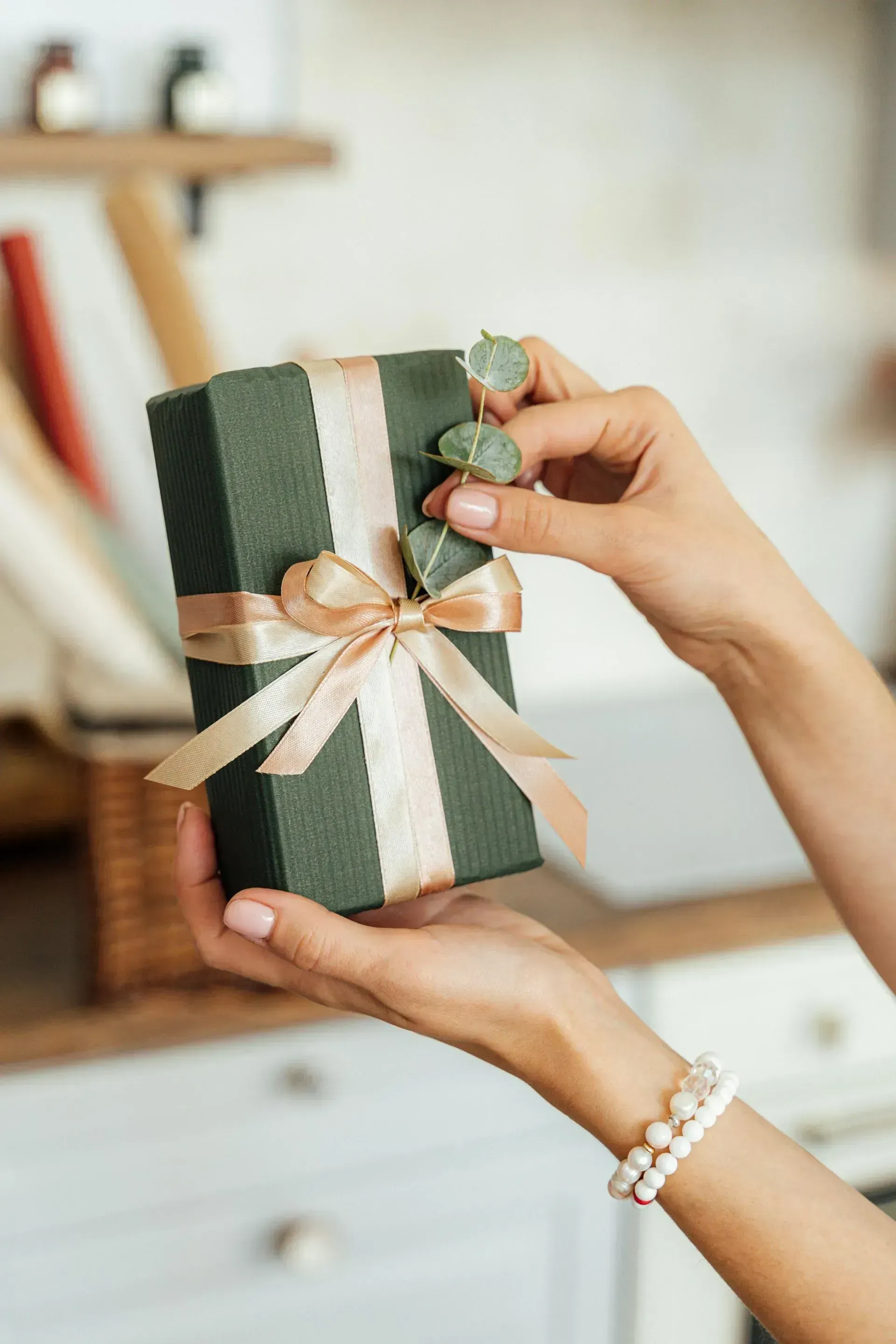 Gold gift card with bow Hands holding a wrapped dark green gift box with a light pink ribbon and sprigs of greenery.
