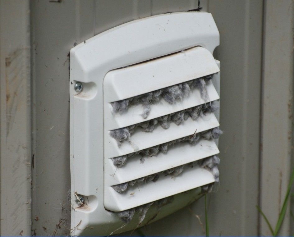 Une grille d'aération blanche pour sècheuse, et obstruée par une importante accumulation de peluches grises.
