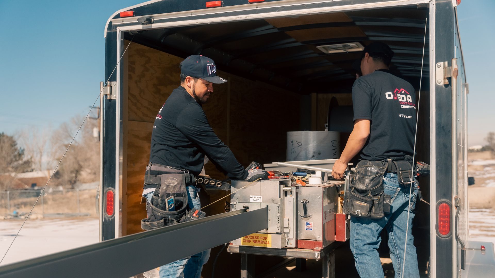 Two men are loading a trailer with a seamless gutter machine in it.