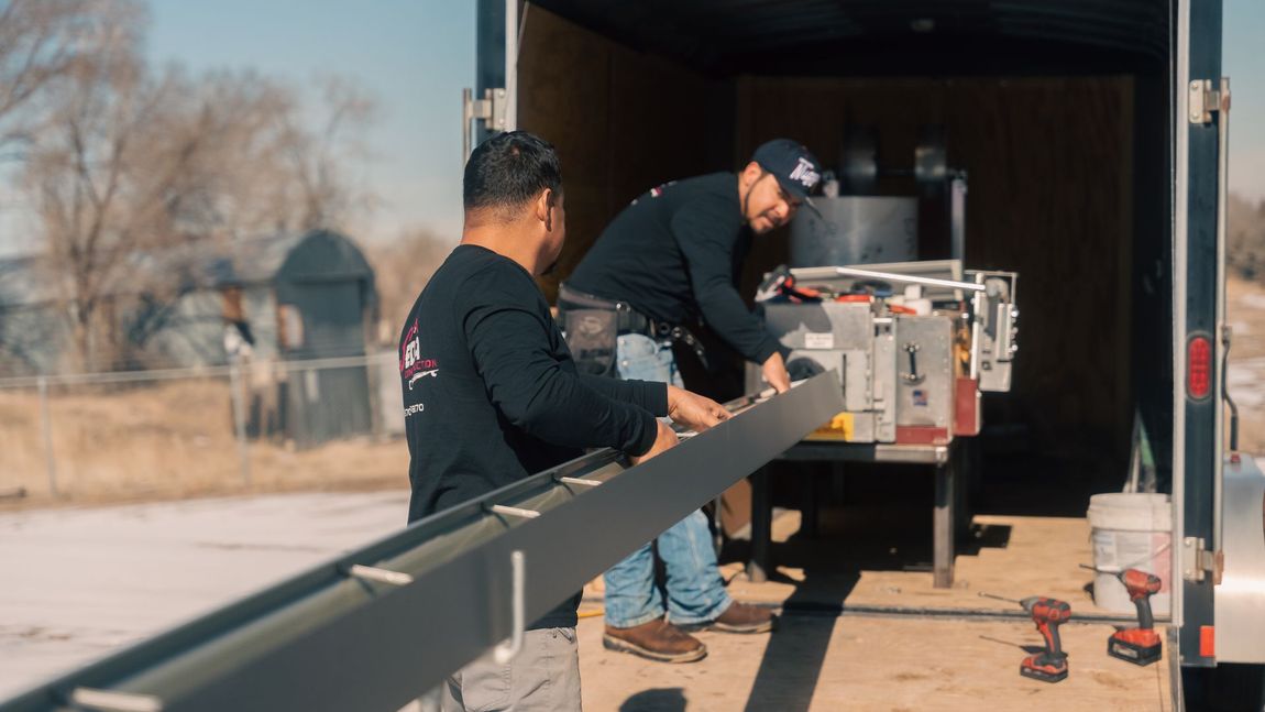 Two men are working on a piece of gutter in front of a trailer.