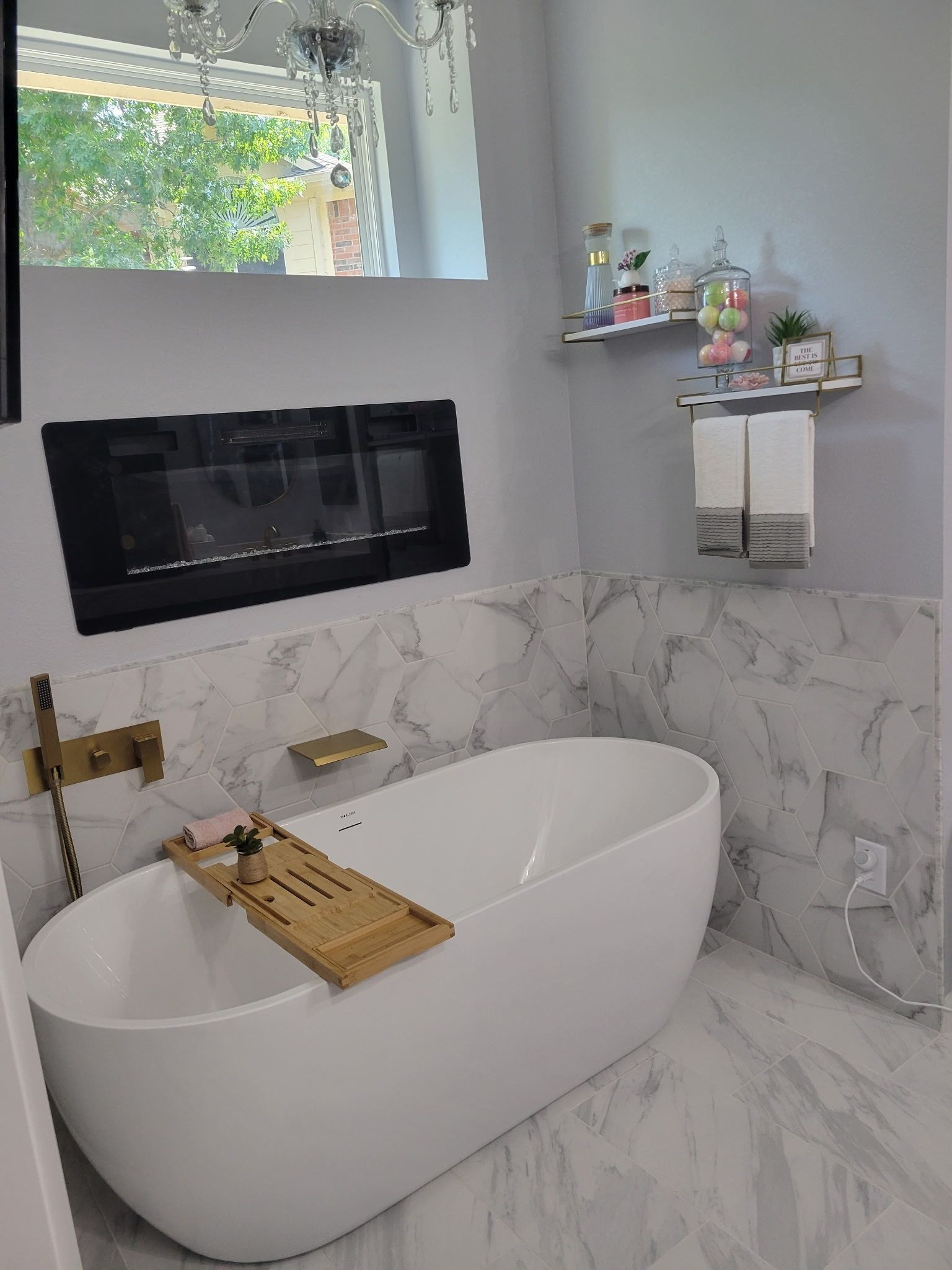 Modern bathroom with a white freestanding tub, marble tile, black fireplace, and window shelf with decor