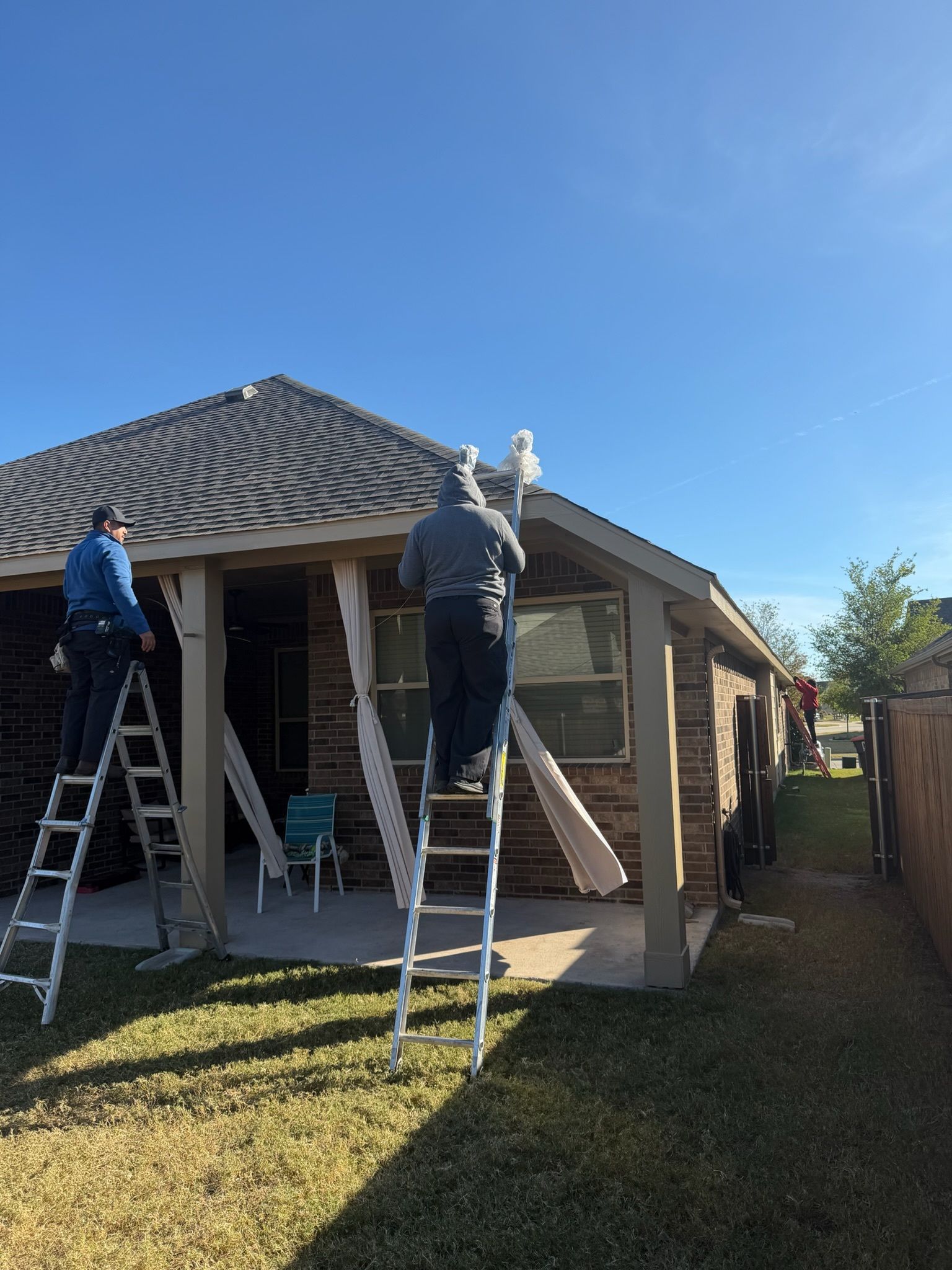 Workers on ladders repairing a house roof and porch under a clear blue sky.