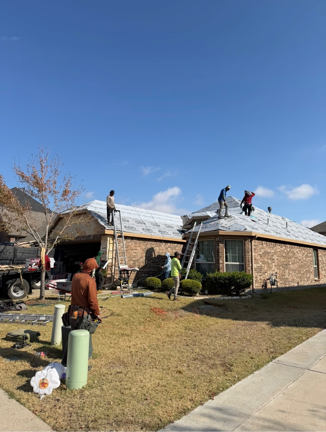 Workers on a brick house roof during repairs, with tools on the lawn and a clear blue sky.