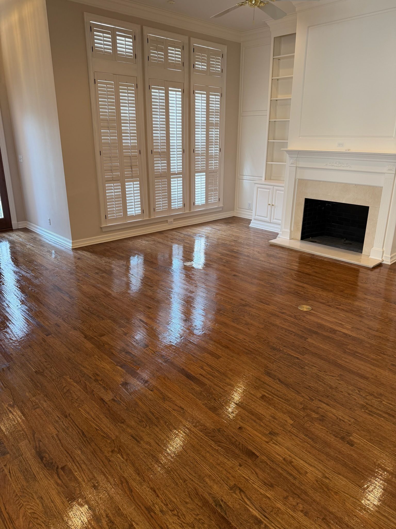 Empty living room with glossy hardwood floors, white shutters, and a fireplace with built-in shelves.