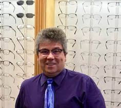 A man in a purple shirt and blue tie is standing in front of a wall of glasses.