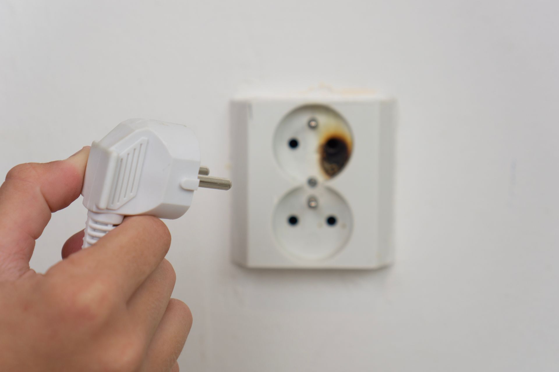 Hand holding a white electrical plug near a damaged wall outlet with burn marks. Hand holding a white electrical plug near a damaged wall outlet with burn marks.