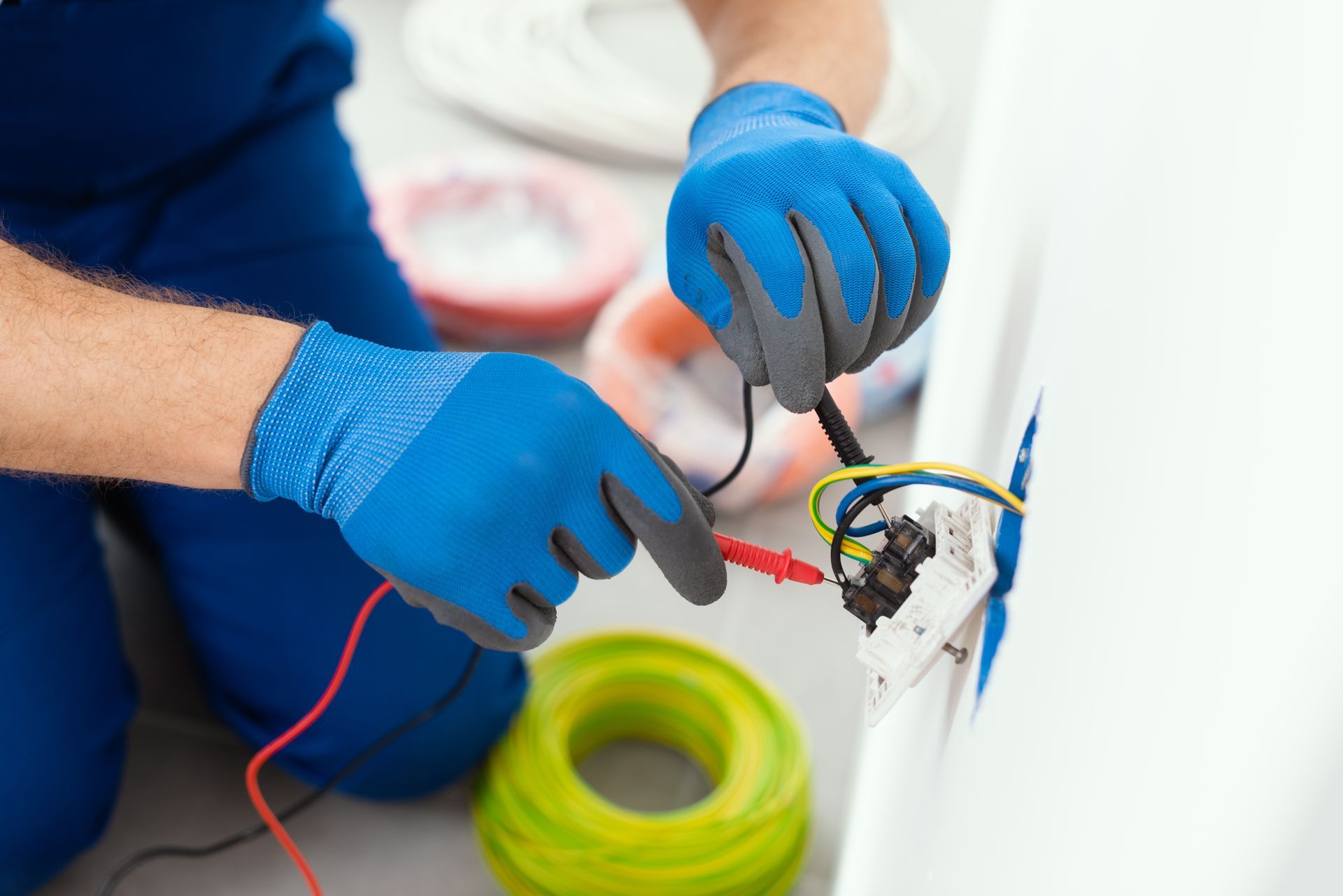 Electrician in blue gloves testing a power outlet with a multimeter; cables and spools visible. Electrician in blue gloves testing a power outlet with a multimeter; cables and spools visible.