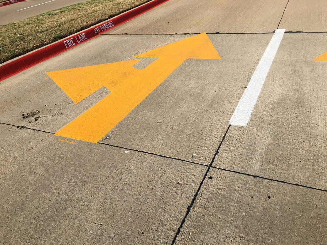Yellow arrow marking on a concrete surface, indicating a direction. Beside a white stripe and red curb.