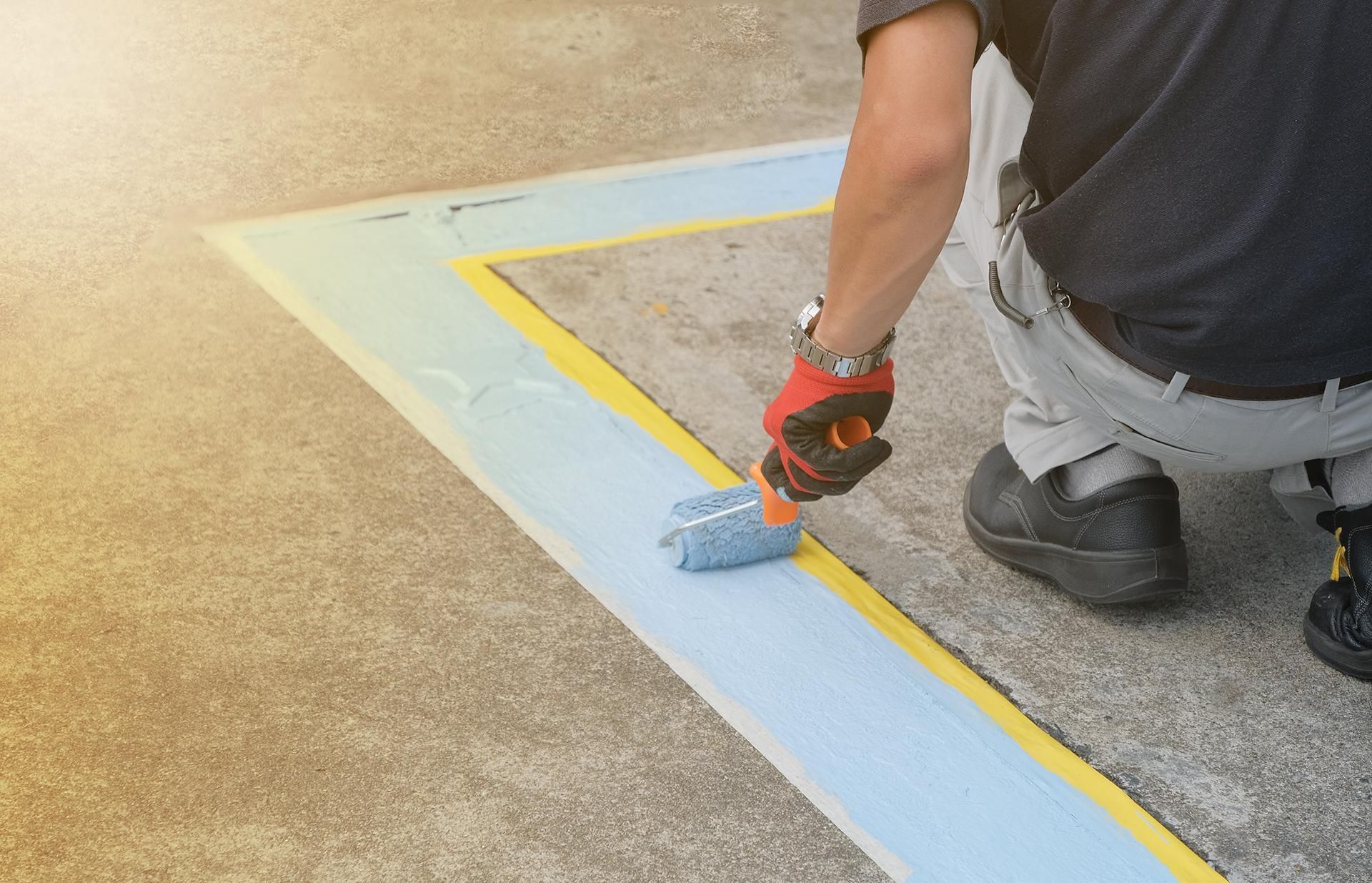 Person painting a blue line inside a yellow-taped border on concrete, using a small roller.