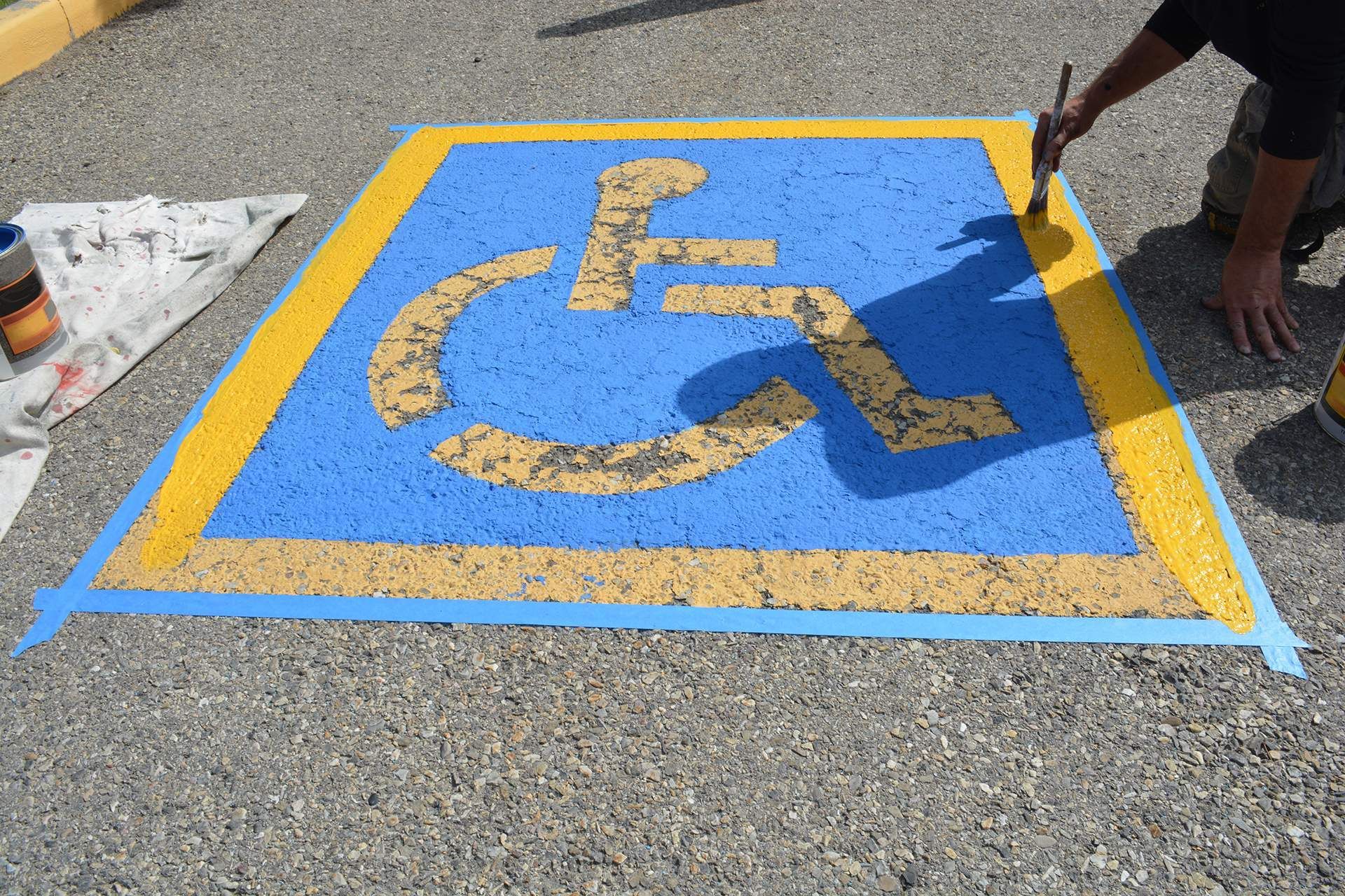 Person painting a blue and yellow handicap parking space symbol on pavement.
