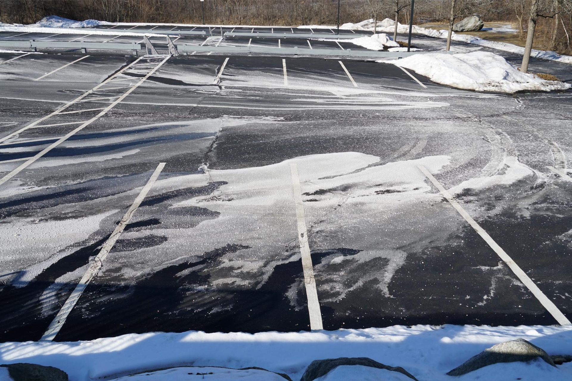 An asphalt parking lot partially covered in patches of snow and ice, featuring painted white parking space lines.