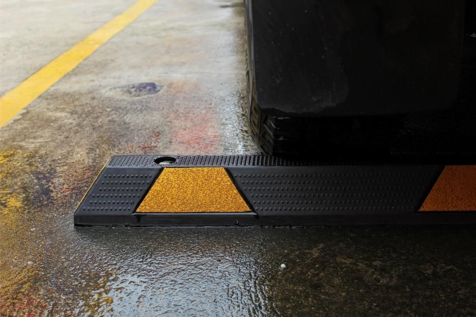 A black parking curb with reflective orange trapezoidal markers, situated on a wet concrete parking space near a car tire.
