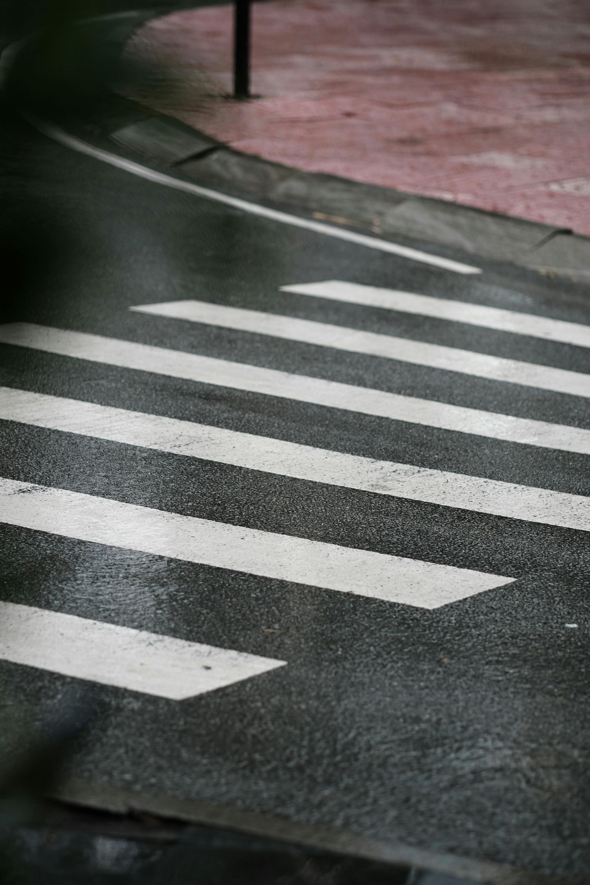 Crosswalk on a wet asphalt road, with white stripes against dark gray. Red sidewalk in background.