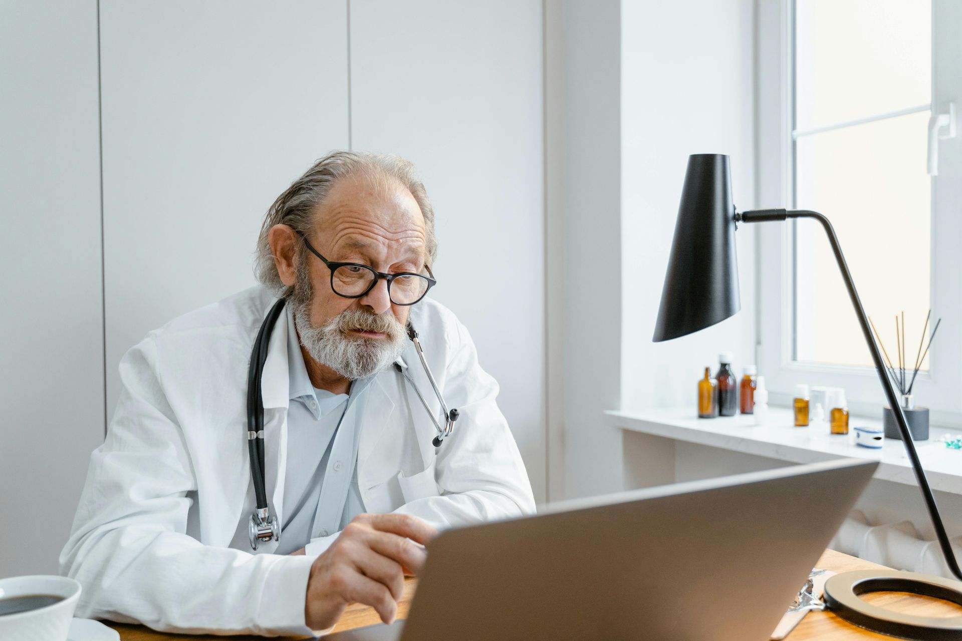 Doctor with glasses and stethoscope looking at laptop, in an office setting.
