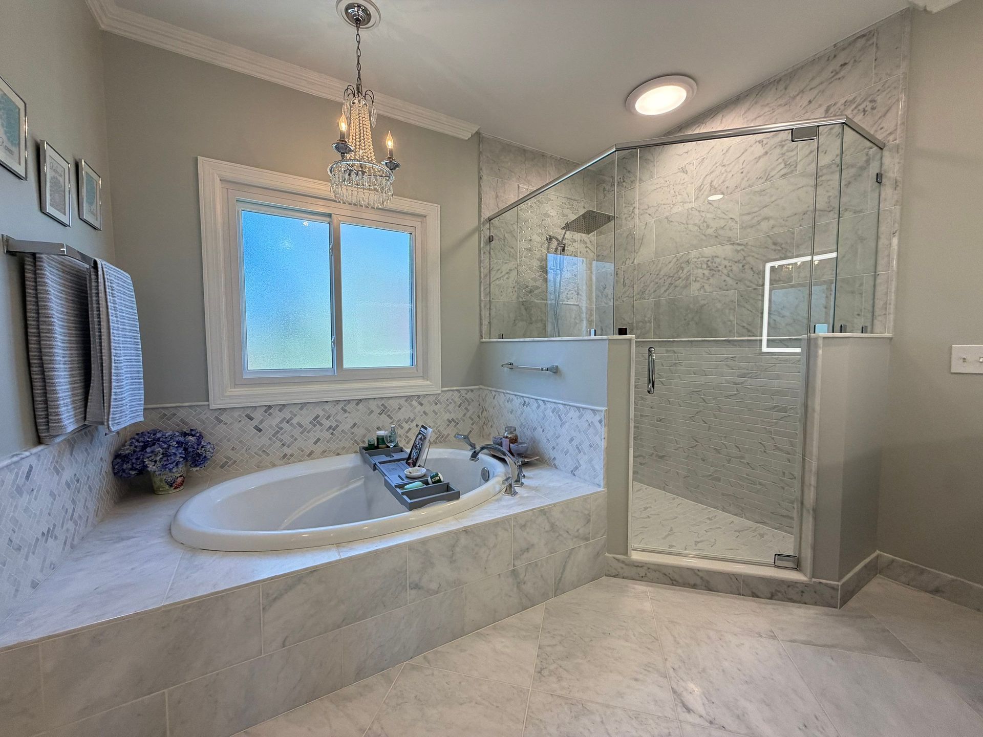 Bright marble bathroom with a soaking tub, glass shower, and window under a chandelier.