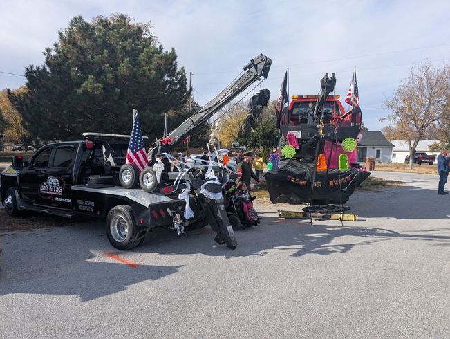 Tow truck loading a decorated float in a street. Crew members, American flag. Sunny day.
