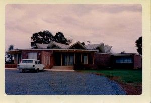 A mid-century modern brick house with a distinctive zigzag roofline and a white station wagon parked in the gravel driveway.