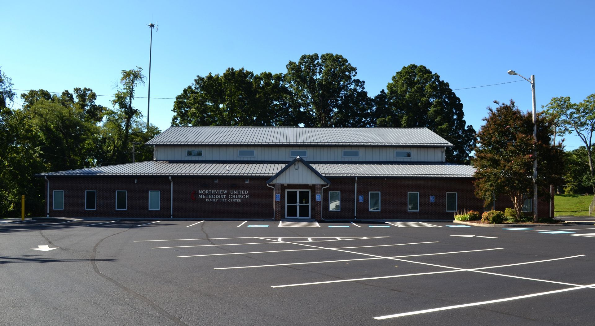 A one-story brick building with a metal roof and large parking lot under a clear blue sky.