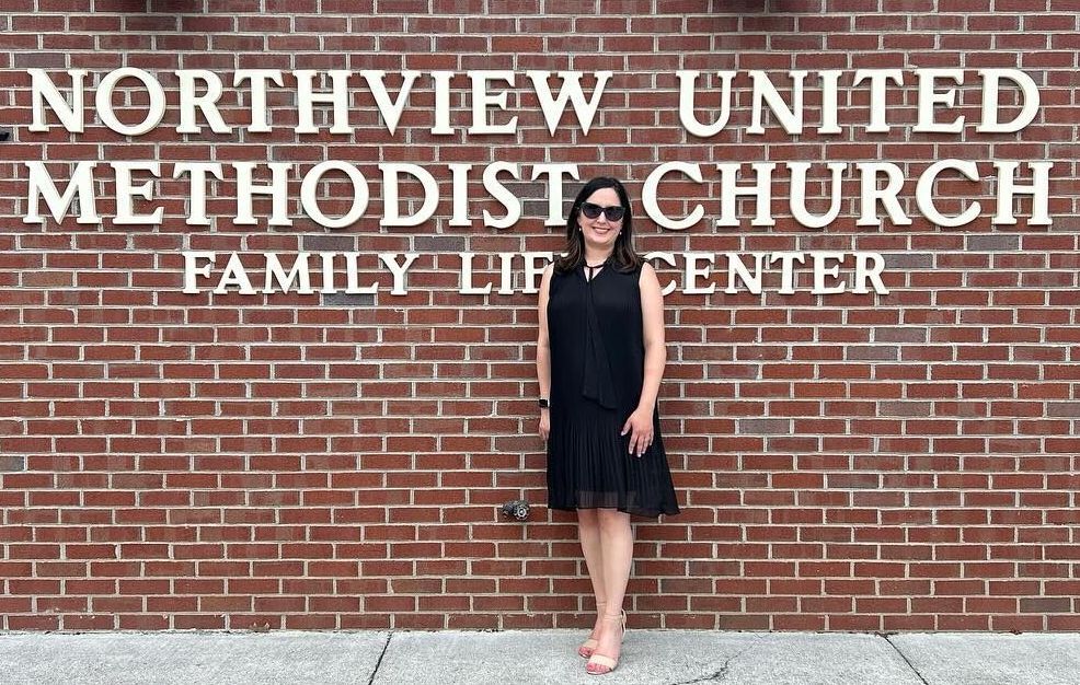 A woman in a black sleeveless dress standing in front of a brick building labeled 