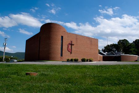 A red brick church building with a cross logo on the front, set against a blue sky with green grass in the foreground.