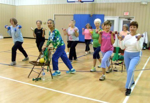 A group of people exercising in a gymnasium, holding small weights and standing in a row with exercise equipment nearby.