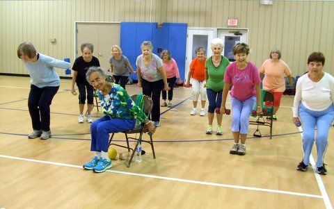 A group of people in an indoor gym performing squats and strength exercises while holding small weights.