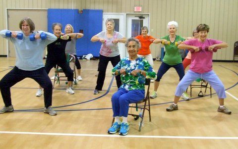 A group of people exercising in a gym, holding hand weights and performing a squat or seated exercise.