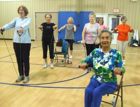 A group of people exercising in a gym, using resistance bands while standing or sitting in chairs.
