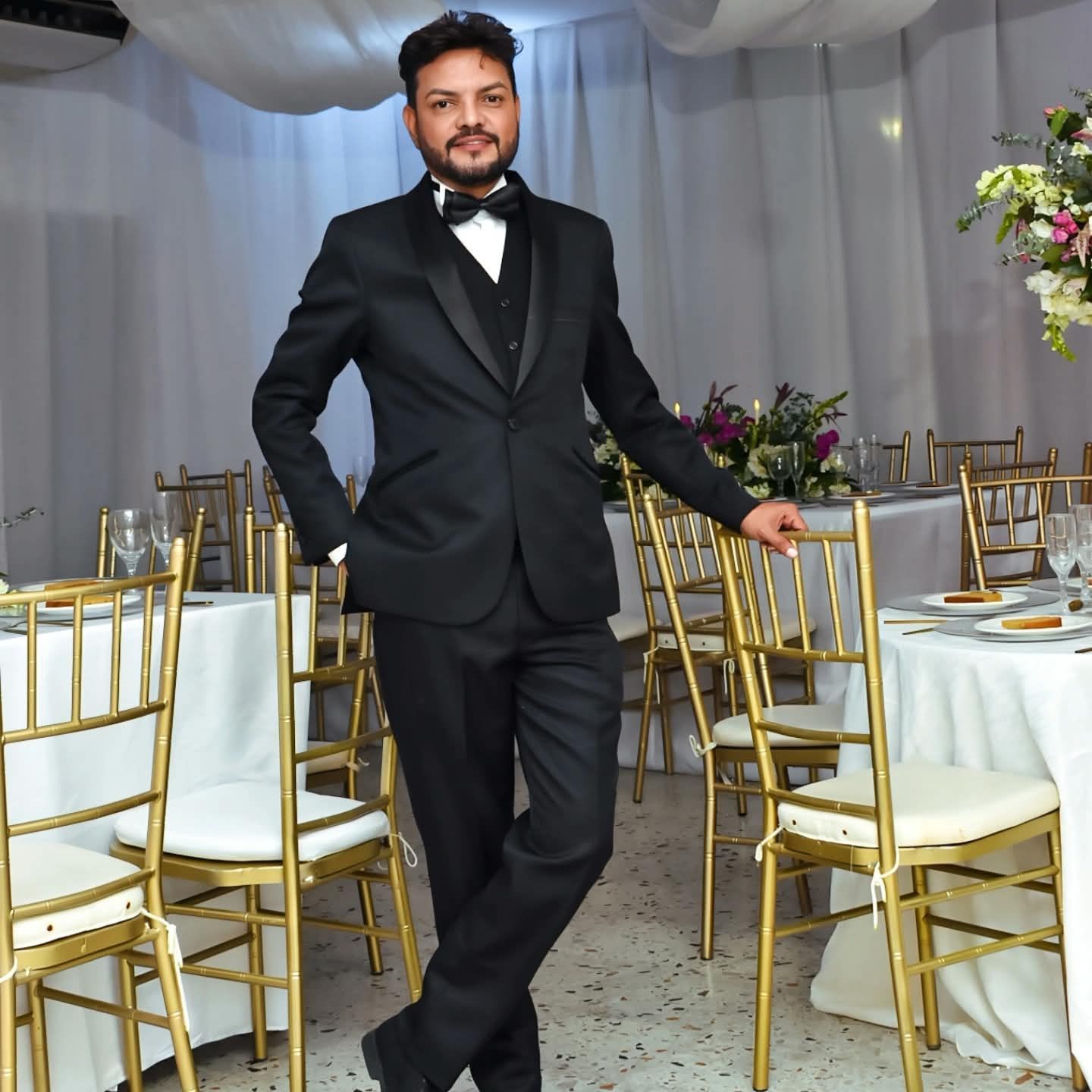 Hombre con esmoquin negro posando entre sillas doradas en un evento formal con mesas preparadas con manteles blancos