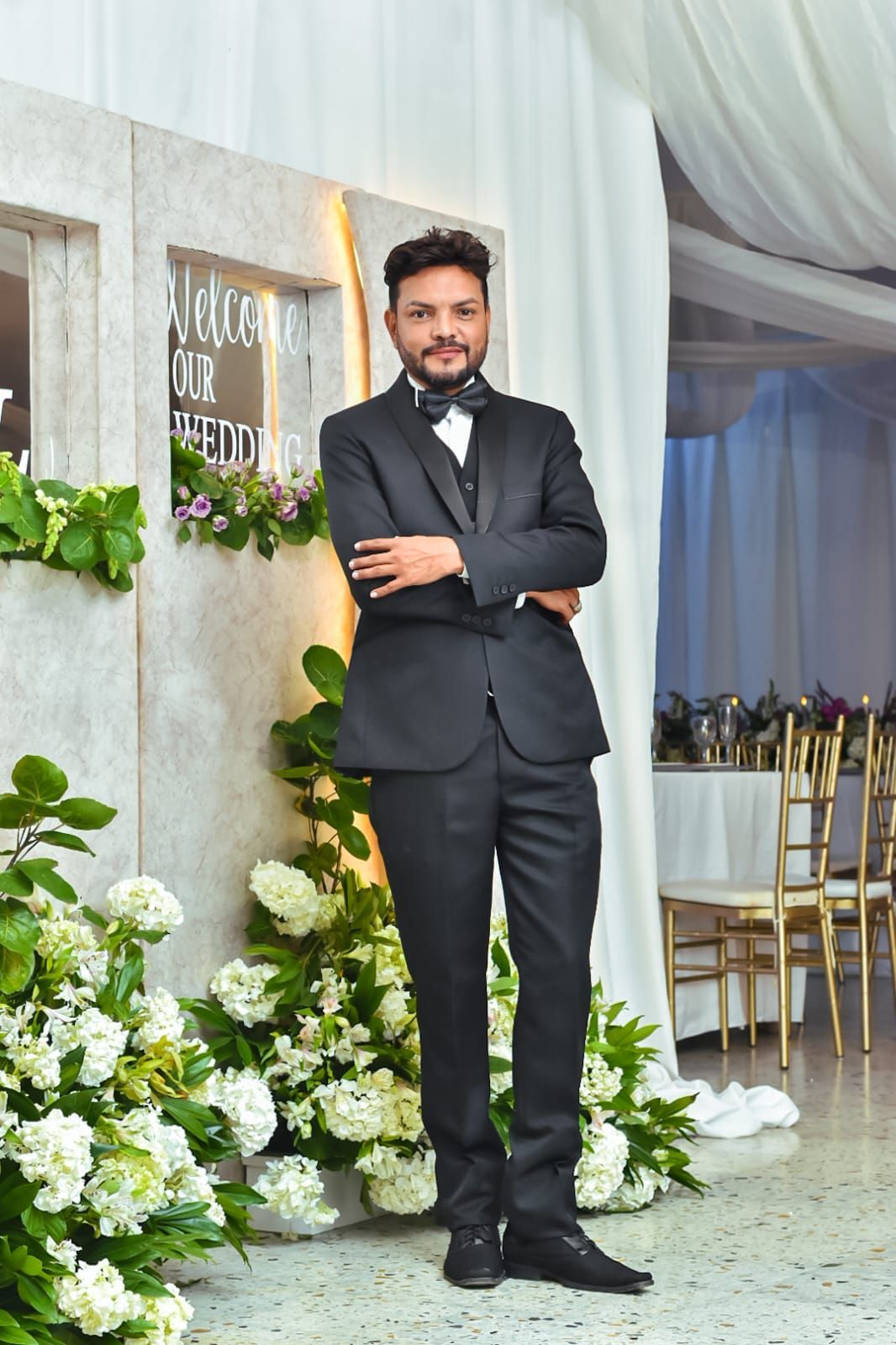 Hombre con traje negro, de pie con los brazos cruzados junto a unos arreglos florales blancos