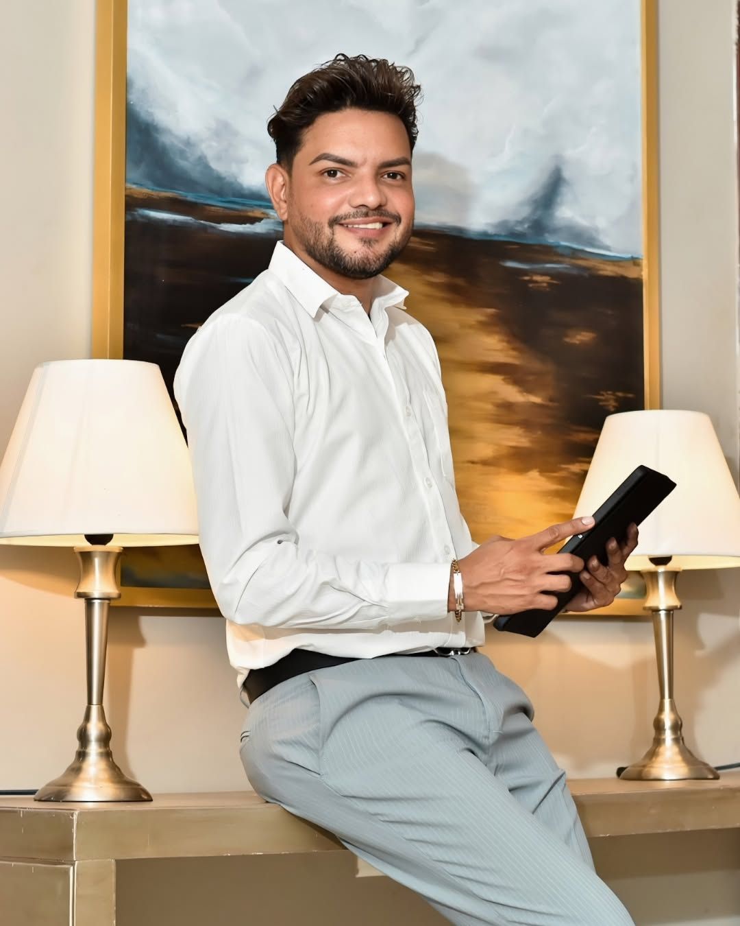Persona sonriente con camisa blanca y pantalones claros sentada en el interior, sosteniendo un teléfono