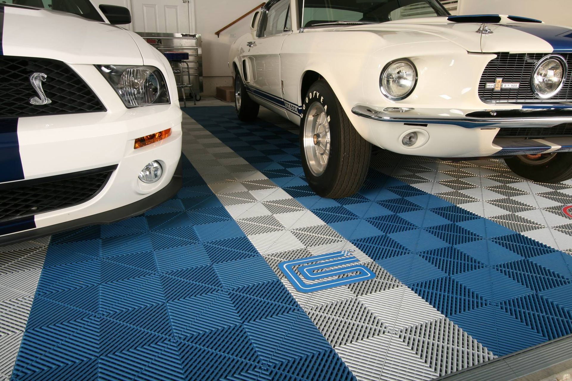 Deux Ford Mustang blanches garées sur le sol carrelé bleu et gris d'un garage.