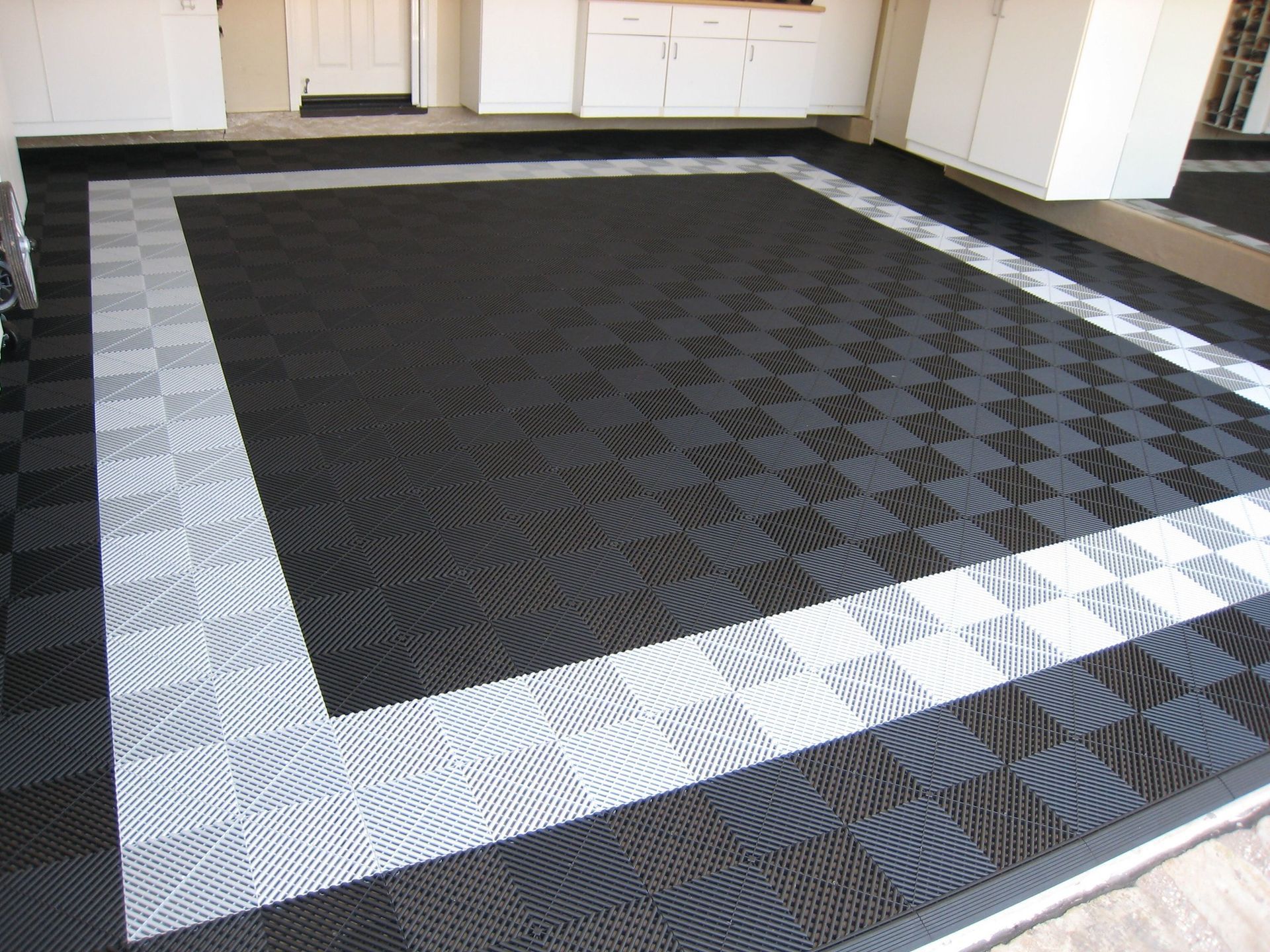 Carrelage de garage emboîtable noir et gris. Bordure blanche, placards blancs et porte visibles.