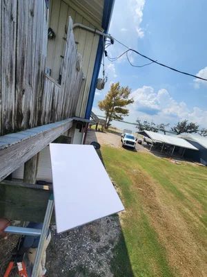 White board leaning against a weathered wooden wall beside a gravel path, with a grassy yard and parked truck beyond.