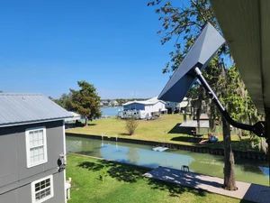 Rural yard with a canal, houses, and a solar panel mounted by a tree under a clear blue sky