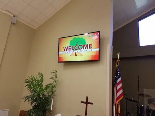 Church sanctuary with a welcome screen, American flag, cross, and potted plant on a beige wall.