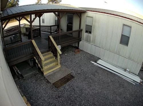 Covered wooden porch entrance with steps and railing beside a white mobile home, gravel yard below