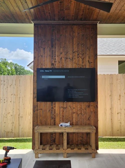 Outdoor TV mounted on a wooden wall under a covered patio, with a wooden console below.
