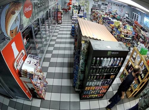 CCTV view of a convenience store aisle with shelves, drinks, snacks, and a checkout counter