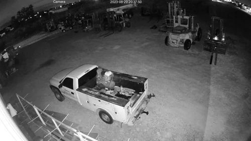 Nighttime security camera view of a white pickup truck parked in a lot near forklifts and equipment.
