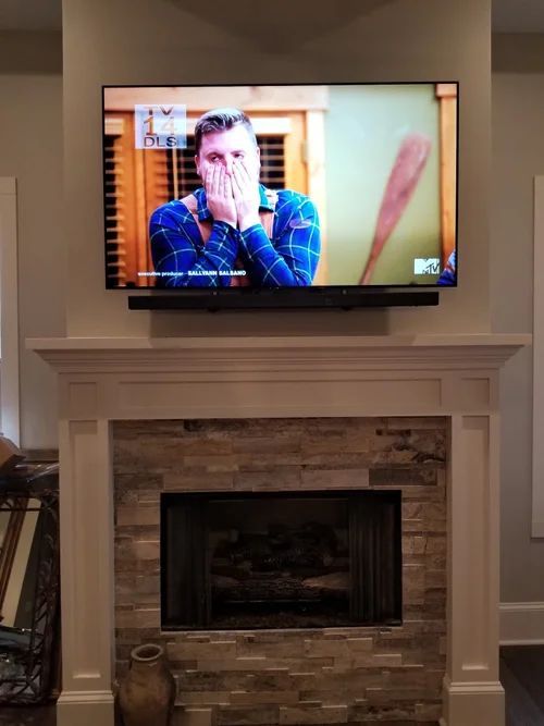 Living room with a wall-mounted TV above a stone fireplace; a man in a blue plaid shirt appears onscreen.