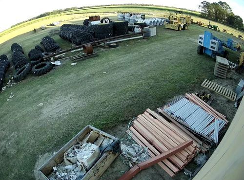 Wide-angle view of a farmyard with stacked tires, scrap wood, pallets, and equipment on grass near fields