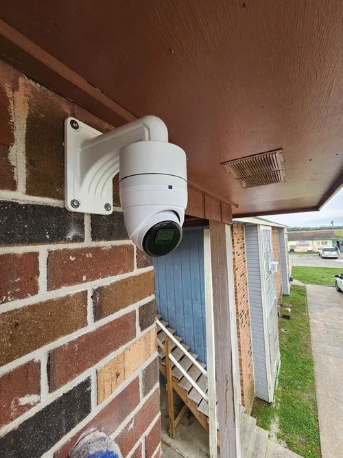White security camera mounted on a brick exterior wall under a porch roof.